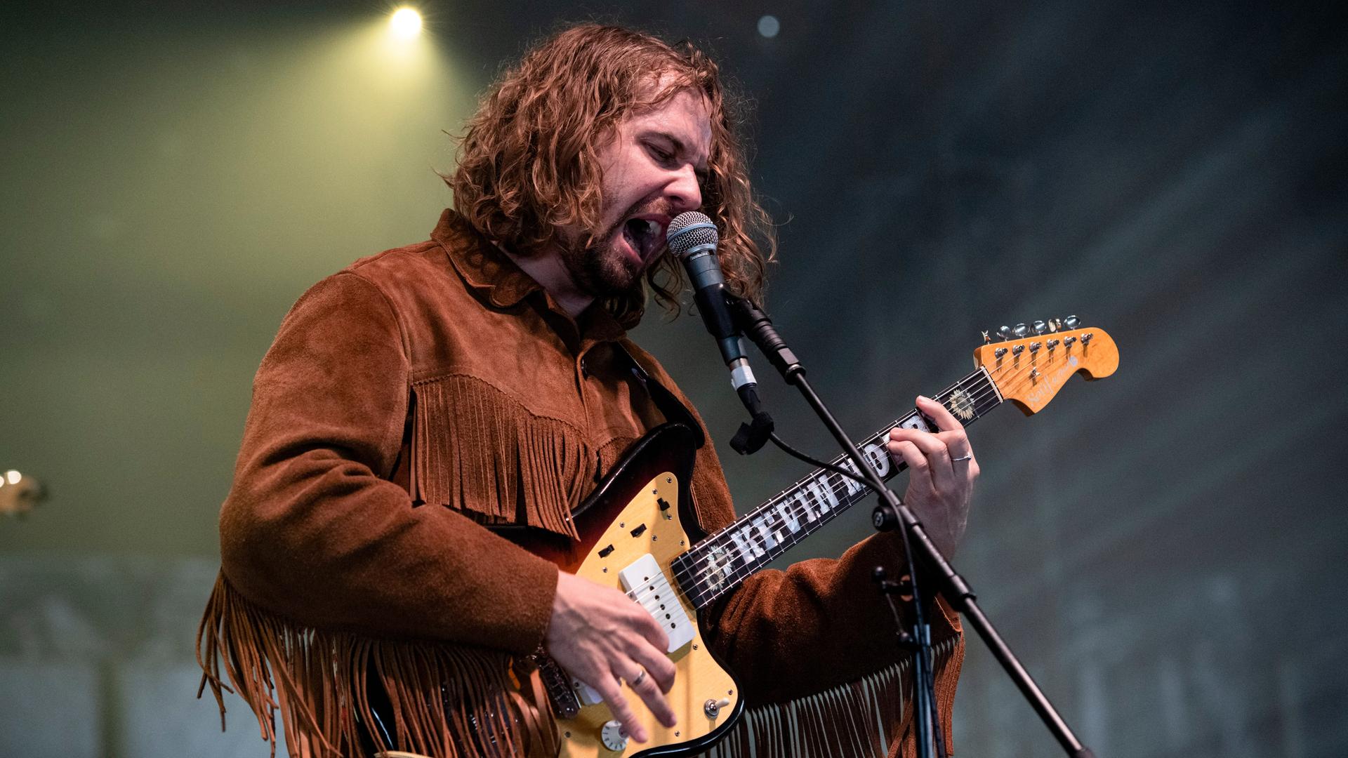 Kevin Morby auf der Bühne. Er trägt eine Jacke mit Fransen und schreit ins Mikrofon, während er Gitarre spielt.