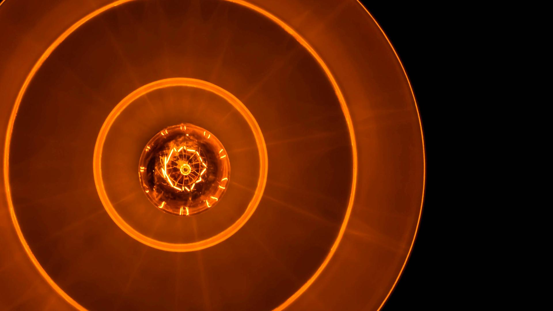 Blick auf eine Glühbirne in tellerförmiger Lampe, die rostrot-orangenes Licht in eine schwarze Umgebung abgiebt.