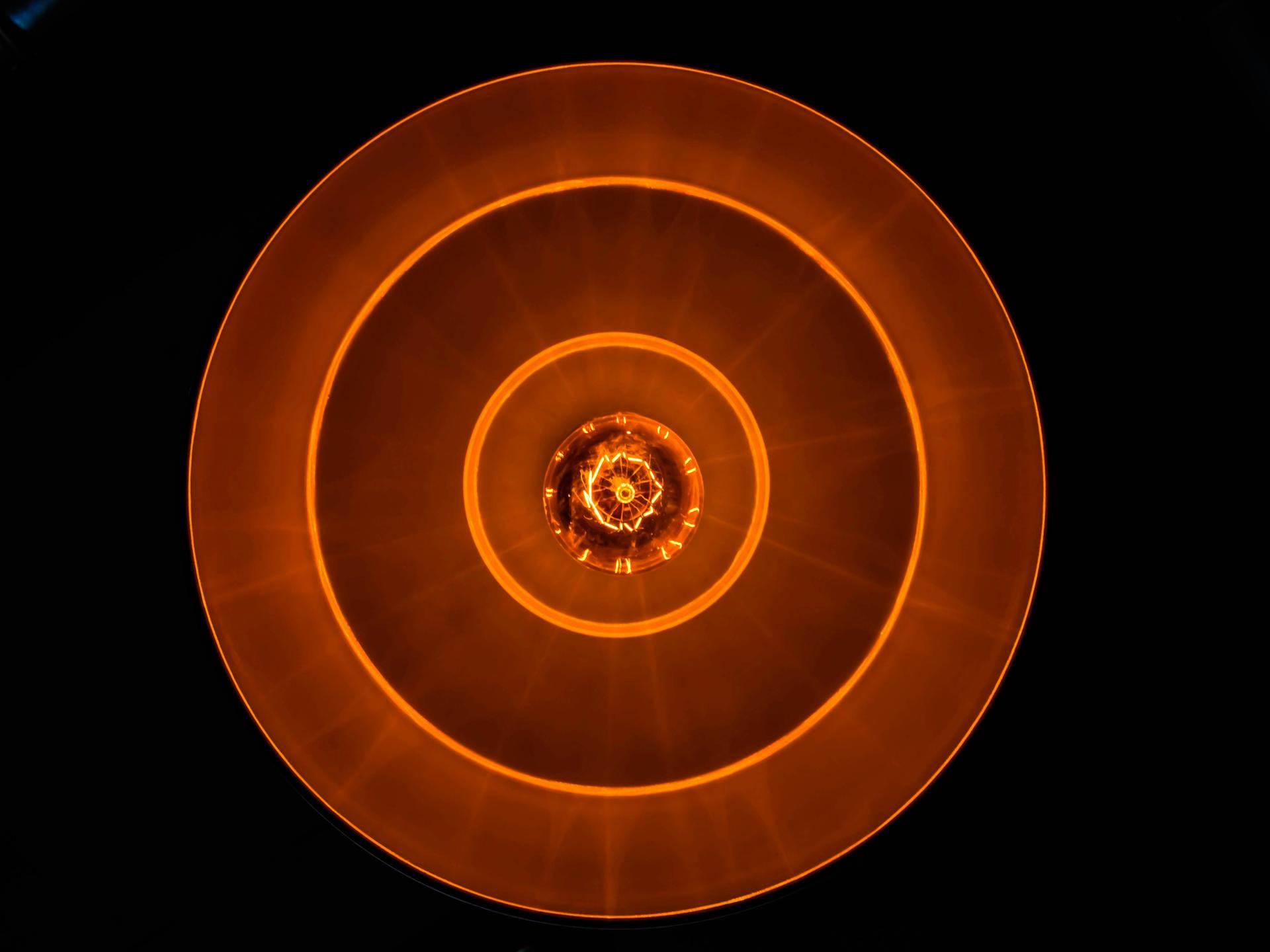 Blick auf eine Glühbirne in tellerförmiger Lampe, die rostrot-orangenes Licht in eine schwarze Umgebung abgiebt.