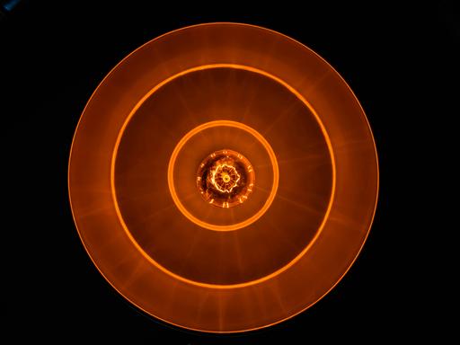 Blick auf eine Glühbirne in tellerförmiger Lampe, die rostrot-orangenes Licht in eine schwarze Umgebung abgiebt.