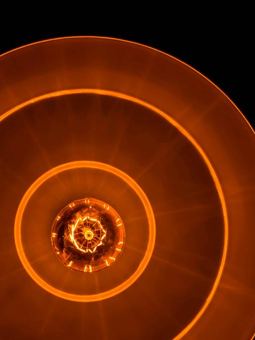 Blick auf eine Glühbirne in tellerförmiger Lampe, die rostrot-orangenes Licht in eine schwarze Umgebung abgiebt.