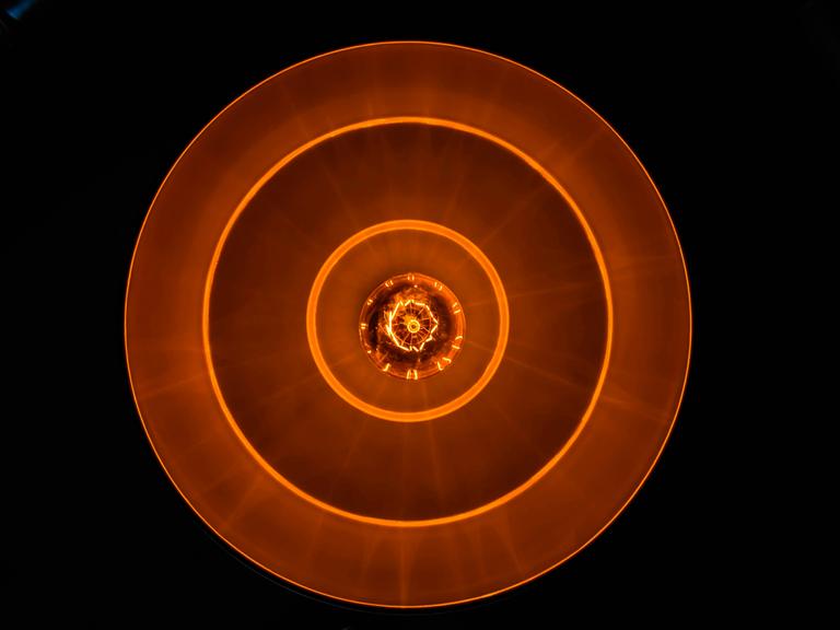 Blick auf eine Glühbirne in tellerförmiger Lampe, die rostrot-orangenes Licht in eine schwarze Umgebung abgiebt.
