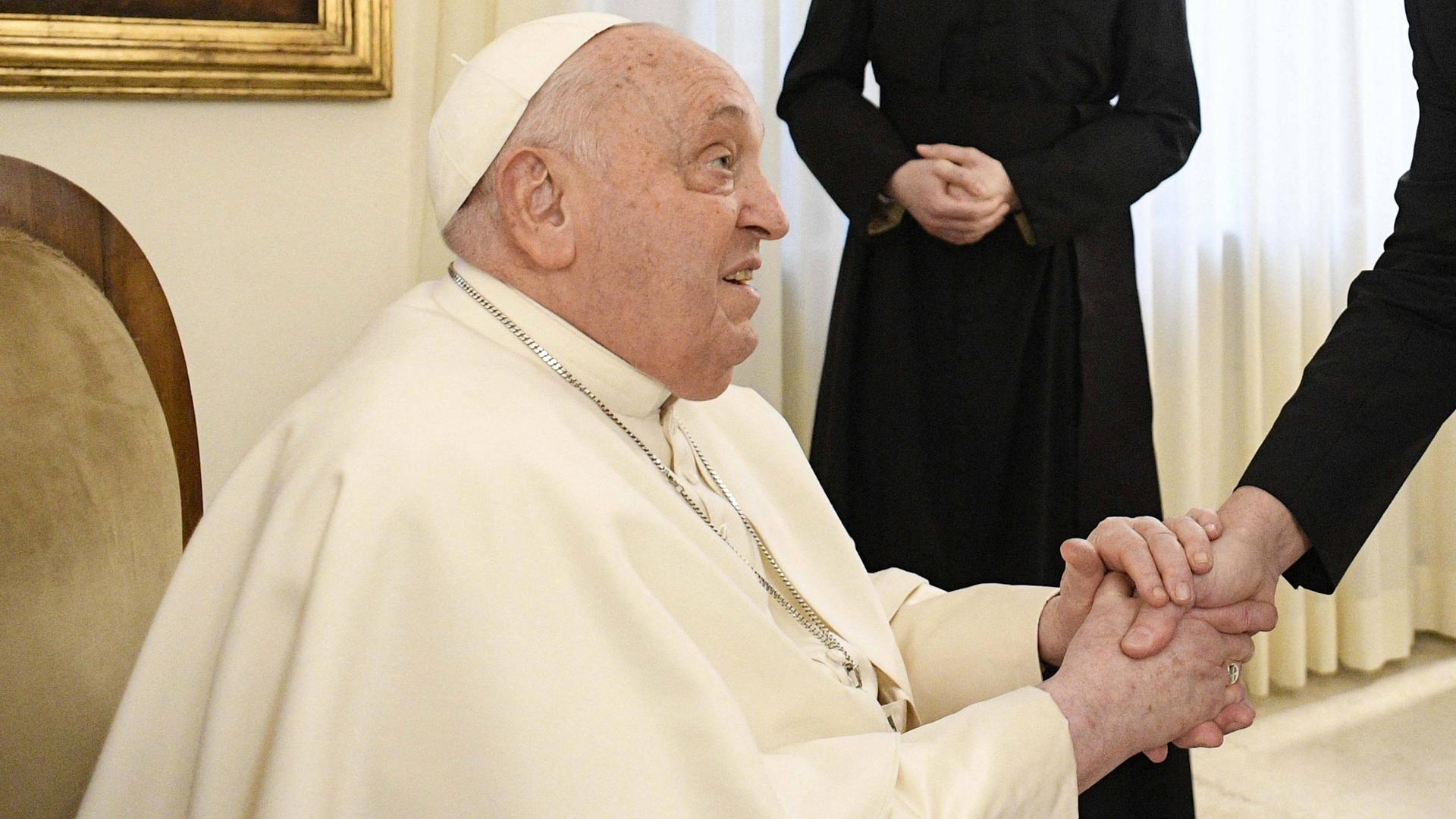 Das Foto zeigt Papst Franzikus. Er sitzt und schüttelt eine Hand.