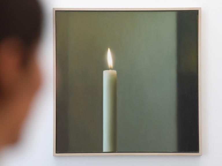 In einer Ausstellung hängt das Bild "Kerze" von Gerhard Richter, links davor sieht man verschwommen eine Frau, die das Bild betrachtet.