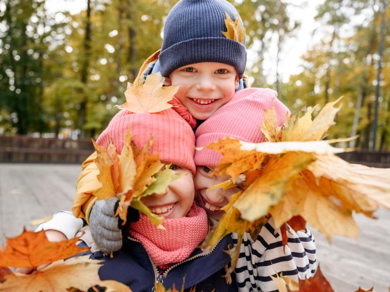 Drei lachende Geschwister spielen mit Herbstlaub. 