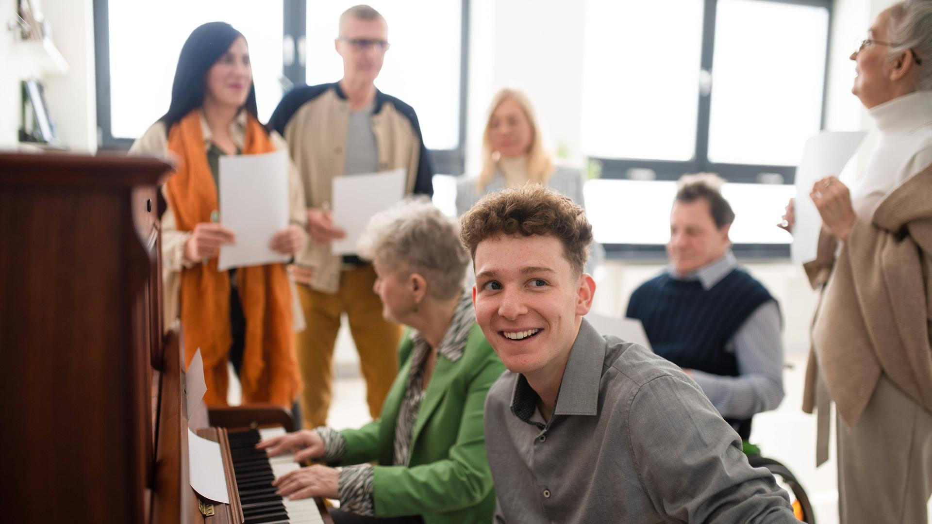 Laienchor bei der Probe. Zwei Personen sitzen am Klavier, andere stehen mit Notenblättern um sie herum.