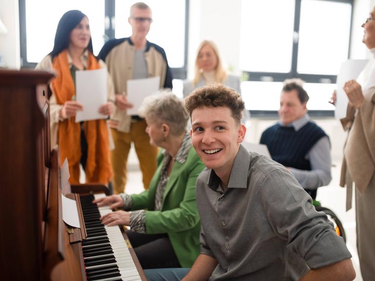 Laienchor bei der Probe. Zwei Personen sitzen am Klavier, andere stehen mit Notenblättern um sie herum.