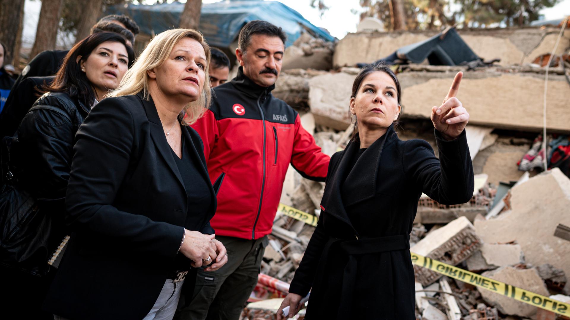Bundesaußenministerin Baerbock und Innenministerin Faeser stehen vor eingestürzten Häusern. Sie werden von einer türkischen Delegation begleitet.