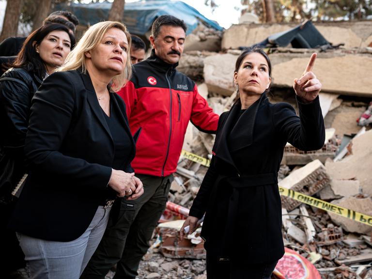 Bundesaußenministerin Baerbock und Innenministerin Faeser stehen vor eingestürzten Häusern. Sie werden von einer türkischen Delegation begleitet.