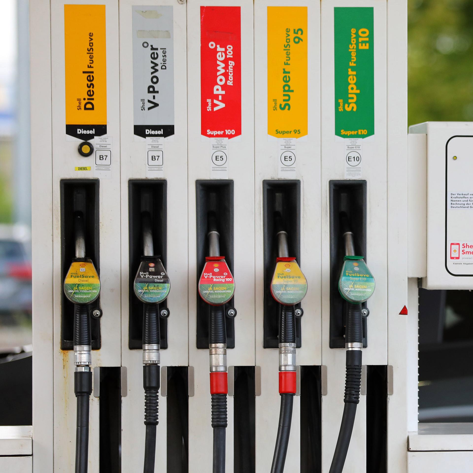 An einer Tankstelle hängen die Zapfhähne für Diesel und Benzin nebeneinander an der Zapfsäule. Im Hintergund steht ein grauer PKW.