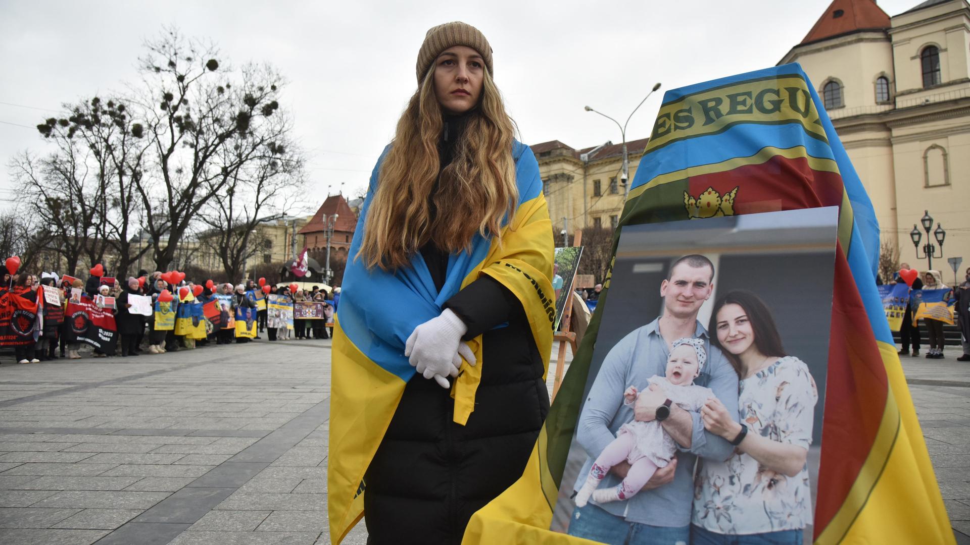 In Lwiw in der Westukraine steht eine Frau mit einer ukrainischen Flagge um die Schultern neben einem großen Bild, das sie mit Mann und Kind zeigt. Sie blickt traurig in die Ferne. 