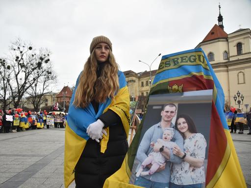 In Lwiw in der Westukraine steht eine Frau mit einer ukrainischen Flagge um die Schultern neben einem großen Bild, das sie mit Mann und Kind zeigt. Sie blickt traurig in die Ferne. 