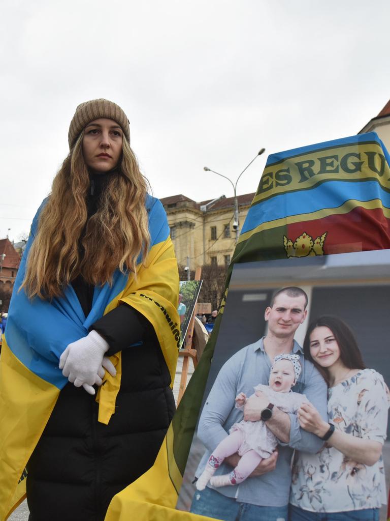 In Lwiw in der Westukraine steht eine Frau mit einer ukrainischen Flagge um die Schultern neben einem großen Bild, das sie mit Mann und Kind zeigt. Sie blickt traurig in die Ferne. 