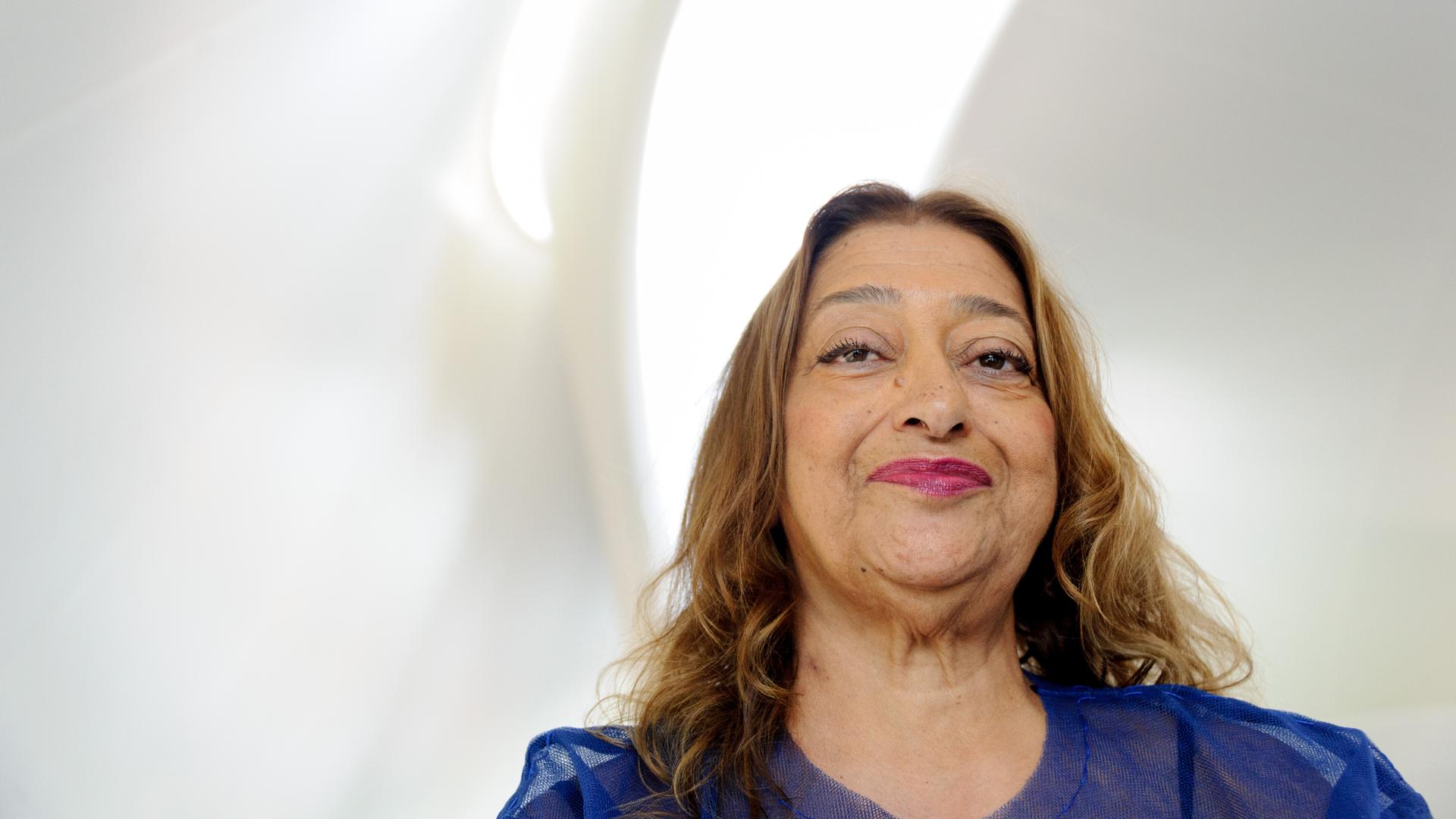 Zaha Hadid lächelt in die Kamera, über ihr fällt Licht durch eine sanft geschwungene Fensteröffnung in den weißen Raum. Zaha Hadid lächelt in die Kamera, über ihr fällt Licht durch eine sanft geschwungene Fensteröffnung in den weißen Raum.