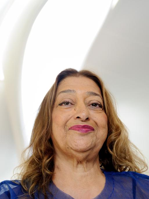 Zaha Hadid lächelt in die Kamera, über ihr fällt Licht durch eine sanft geschwungene Fensteröffnung in den weißen Raum.