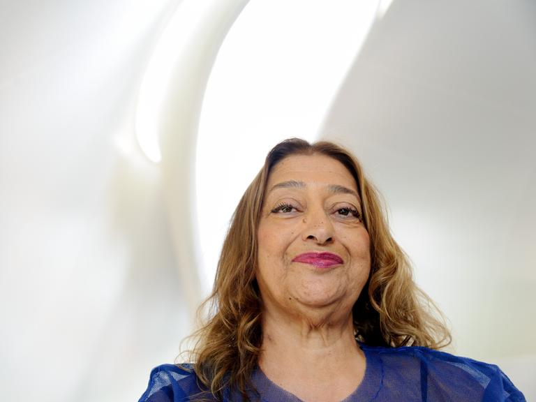 Zaha Hadid lächelt in die Kamera, über ihr fällt Licht durch eine sanft geschwungene Fensteröffnung in den weißen Raum.