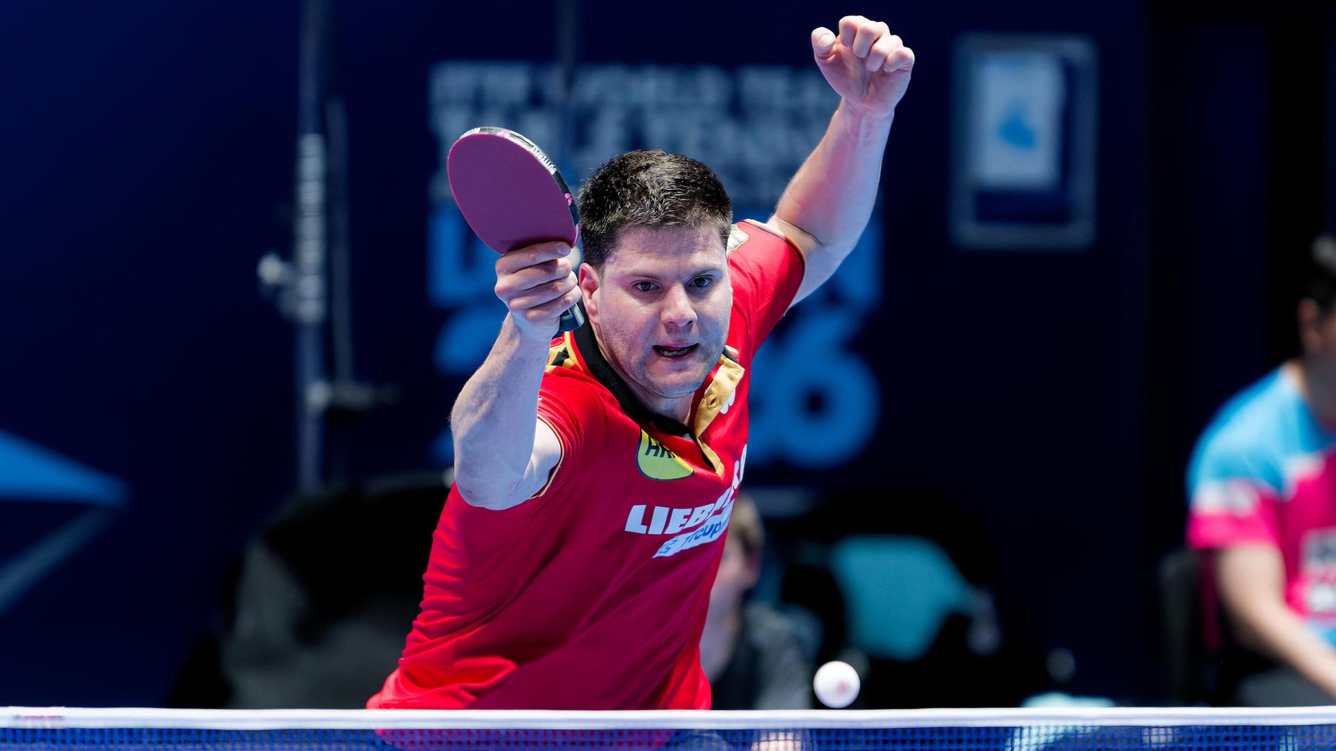 Der Tischtennis-Spieler Dimitrij Ovtcharov hält beide Arme angewinkelt in die Lauft. In der rechten Hand hält er den Schläger. Er blickt konzentriert auf den Tischtennis-Ball vor ihm in der Luft. Der Ball scheint über das Netz zu fliegen. 