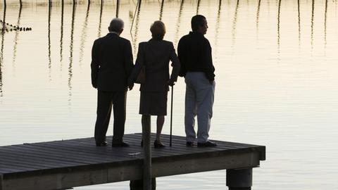Nachdem ein Verständnis aller füreinander erreicht ist, findet die Seele der Mutter ihre letzte Ruhe.. Zu sehen: Zwei Männer und eine Frau stehen auf einem Steg an einem See bei Sonnenuntergang. 