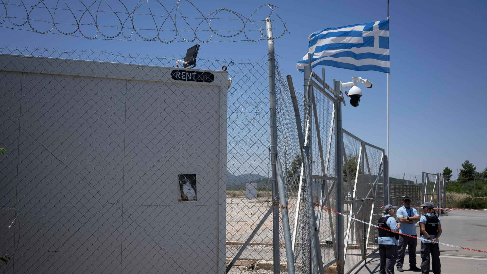 Die griechische Flagge weht am Zaun eines Aufnahmezentrums für Flüchtlinge und Migranten. Davor stehen Sicherheitsleute.