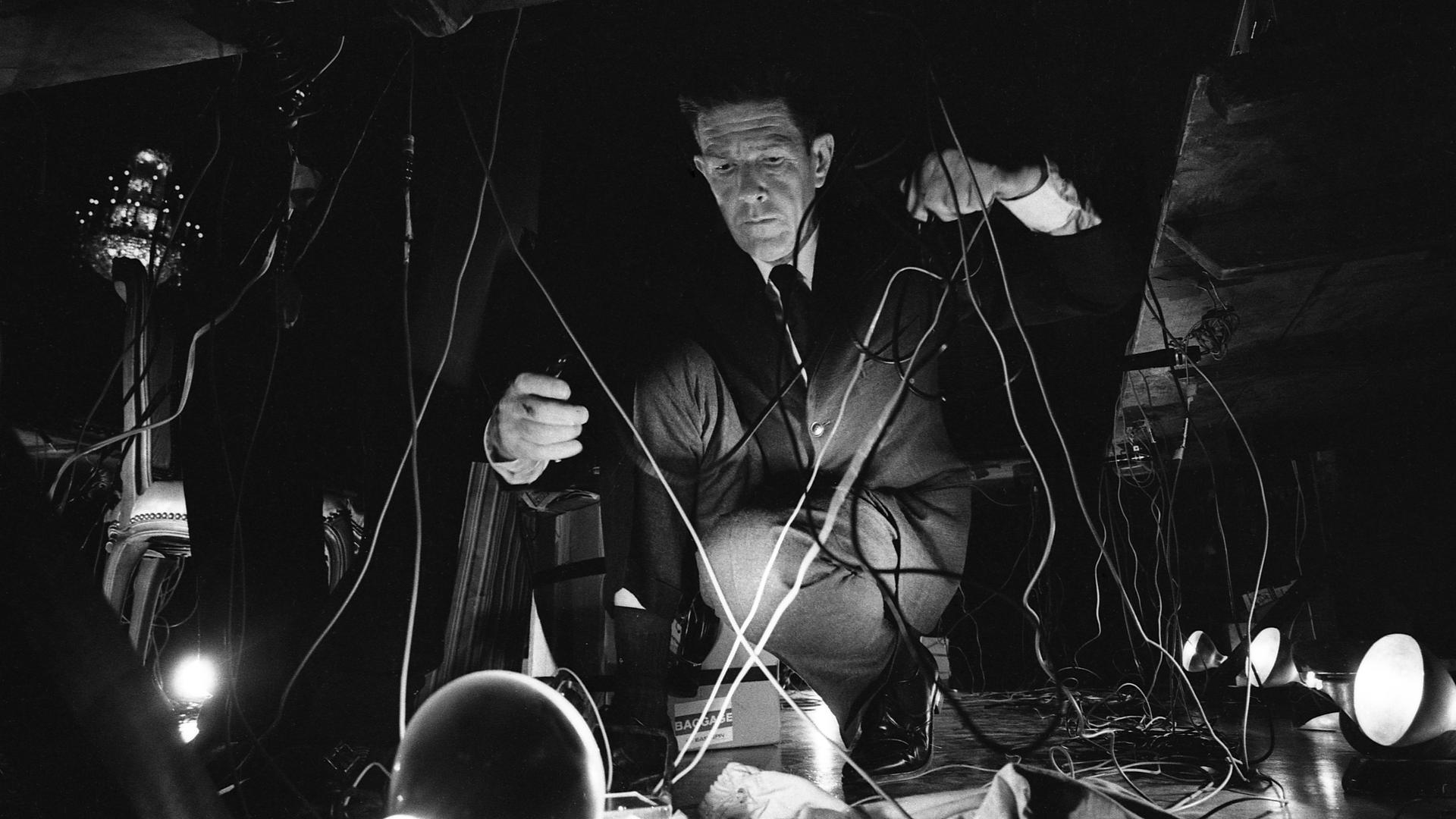 John Cage hockt während eines Konzerts auf dem Boden und hantiert mit Kabeln.