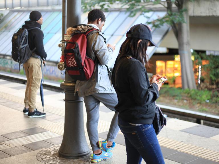 Drei Personen stehen an einem Bahnsteig. Zwei von ihnen schauen auf ihre Smartphones.