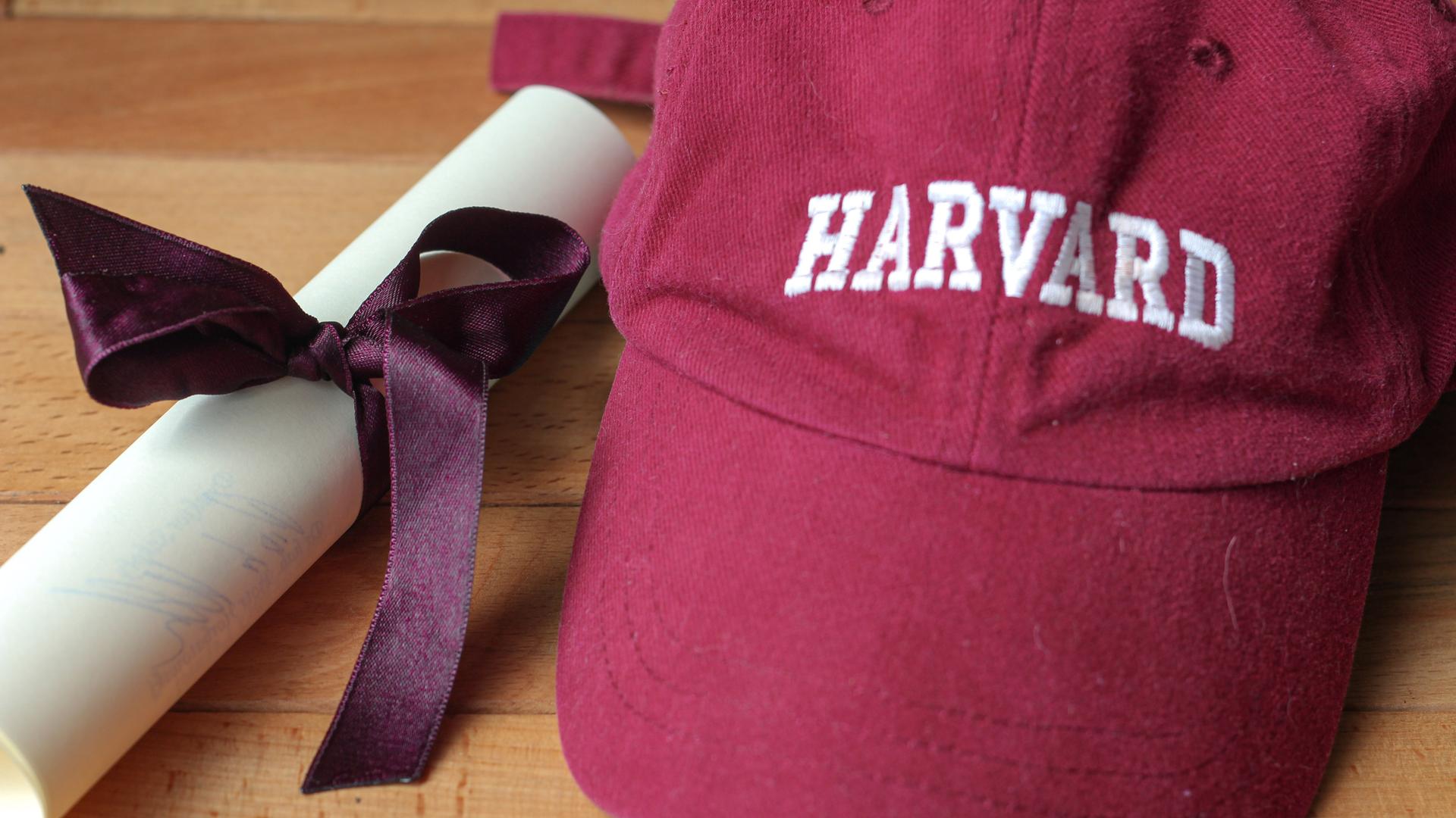 Eine bordeauxfarbene Baseballkappe mit Harvard-Logo und einem eingerollten Dokument, das von einer violetten Schleife zusammengehalten wird.