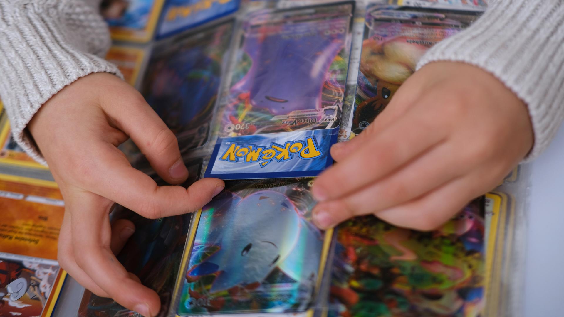 Man sieht einige Pokemon-Karten in einem Sammelalbum und Kinderhände, die eine Karte halten.
