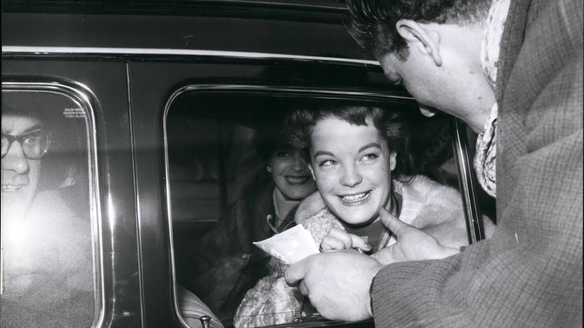 Romy Schneider. Eine junge Frau sitzt in einem Auto und wird noch einem Mann angesprochen.