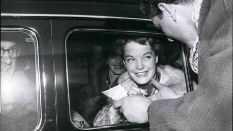 Romy Schneider. Eine junge Frau sitzt in einem Auto und wird noch einem Mann angesprochen.