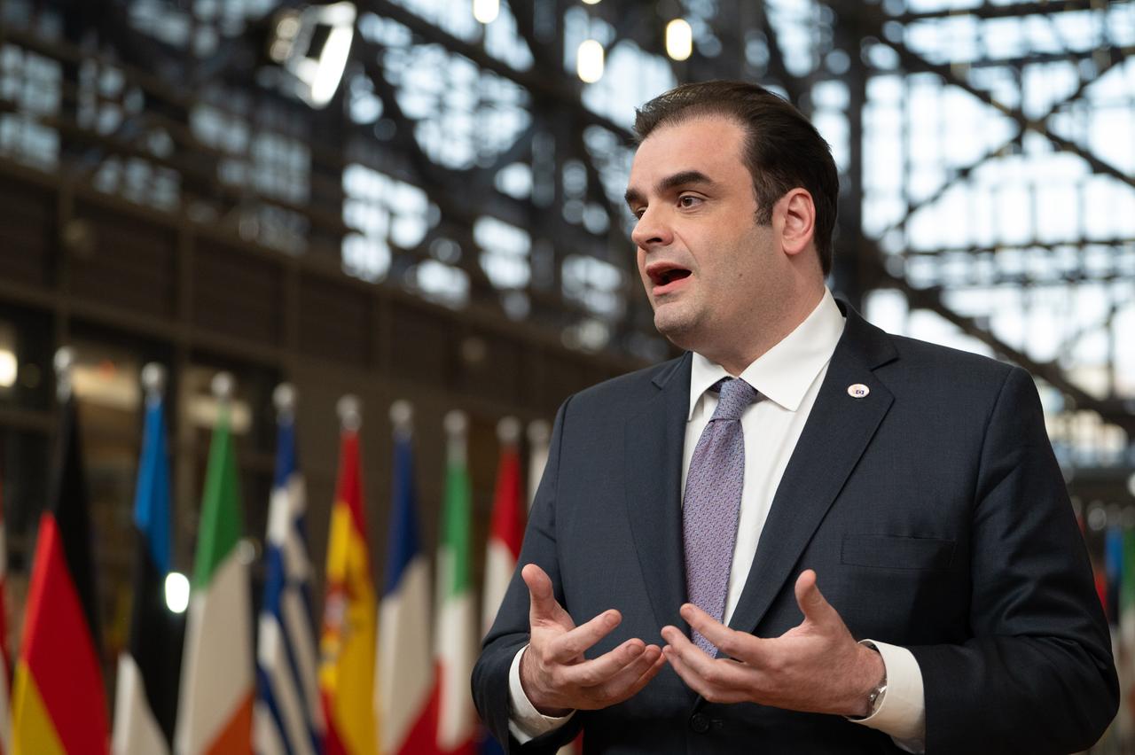 Pierrakakis steht in einem Gebäude, sagt etwas und gestikuliert mit beiden Händen. Dahinter unscharf Flaggen von EU-Staaten.