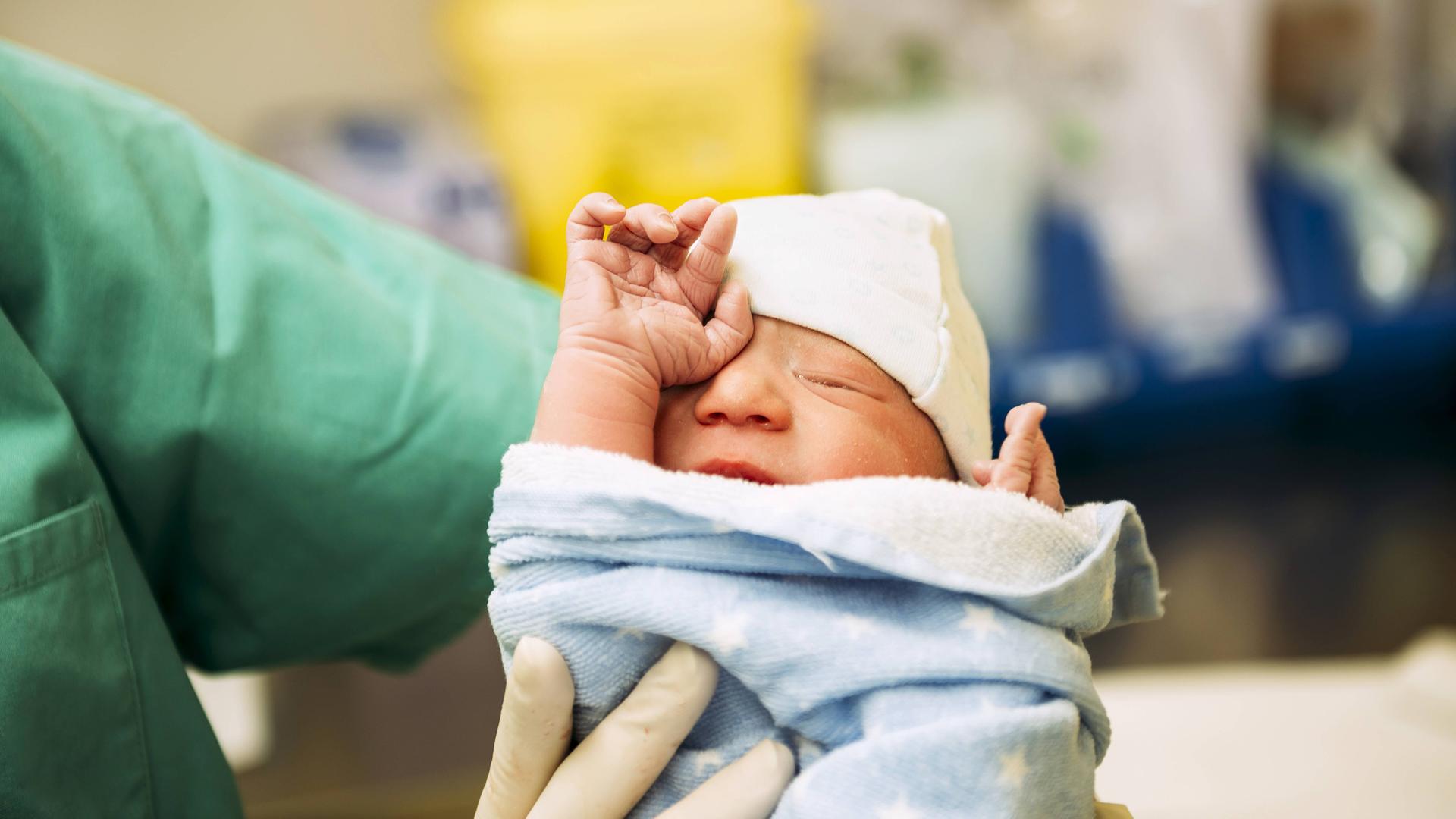 Ein neugeborenes Baby, eingewickelt in ein Handtuch, gehalten von einer Ärztin im Kreißsaal.