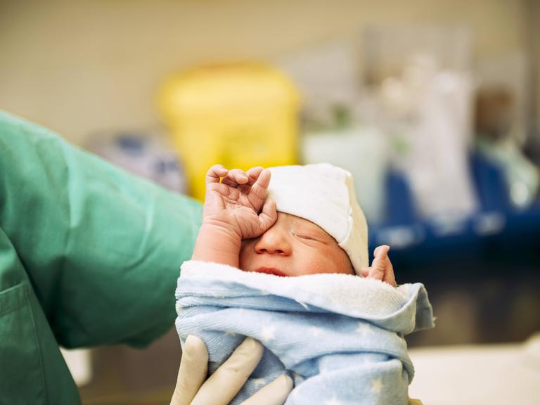 Ein neugeborenes Baby, eingewickelt in ein Handtuch, gehalten von einer Ärztin im Kreißsaal.