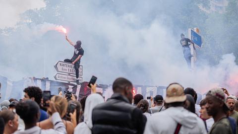 Demonstrationen säumen die Straßen anläßlich des Tods des 17-jährigen Nahel, der bei einer Verkehrskontrolle am Rande von Paris von einem Polizisten erschossen wurde. Auf einem Straßenschild Richtung Nanterre sitzt ein vermummter Mann mit einem Bengalo.