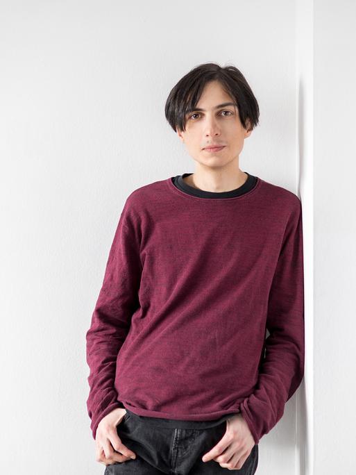 Elias Hirschl lehnt sich in einem Raum voller weißer Wände an eine. Er trägt ein weinrotes Sweatshirt und eine dunkle Hose. Sein schwarzen Haare trägt er mit Mittelscheitel.