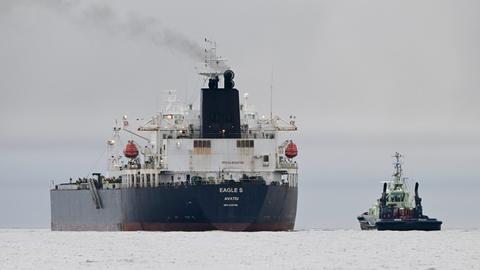 Man sieht den Tanker von hinten mit rauchendem Schlot in der Ferne im Meer, daneben ein kleinerer Schlepper.