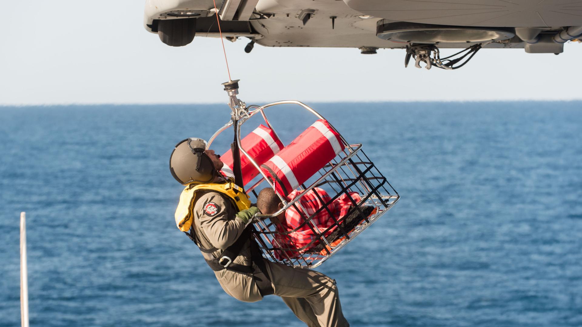 Das Foto zeigt einen Hubschrauber der italienischen Küstenwache über dem Mittelmeer, der einen Korb mit einem Kind und einen Soldaten an einem Seil nach oben zieht.