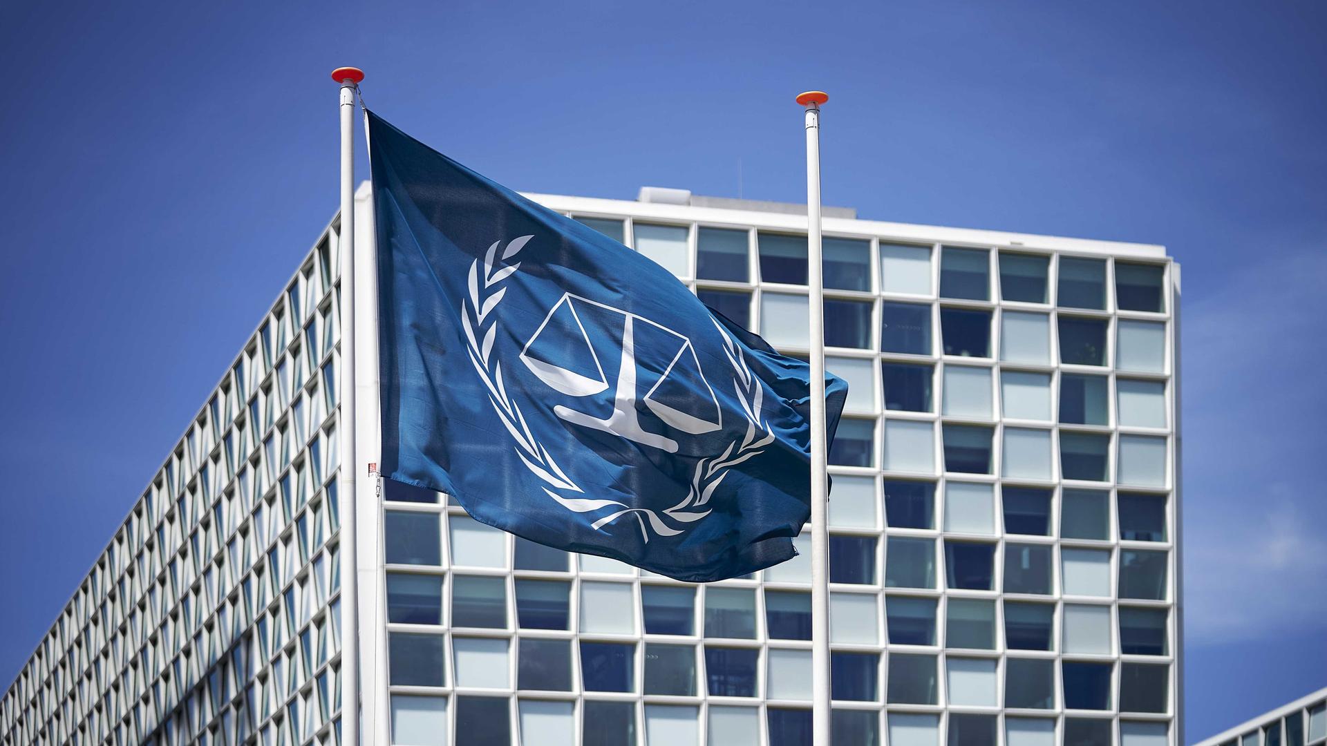 Außenansicht des Internationalen Strafgerichtshofs (IStGH). Davor weht die Flagge des IStGH, eine Waage mit zwei Waagschalen, die von zwei Olivenzweigen umschlossen wird.