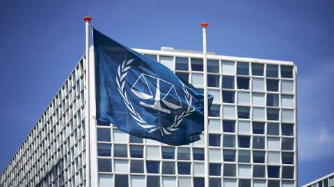 Außenansicht des Internationalen Strafgerichtshofs (IStGH). Davor weht die Flagge des IStGH, eine Waage mit zwei Waagschalen, die von zwei Olivenzweigen umschlossen wird.