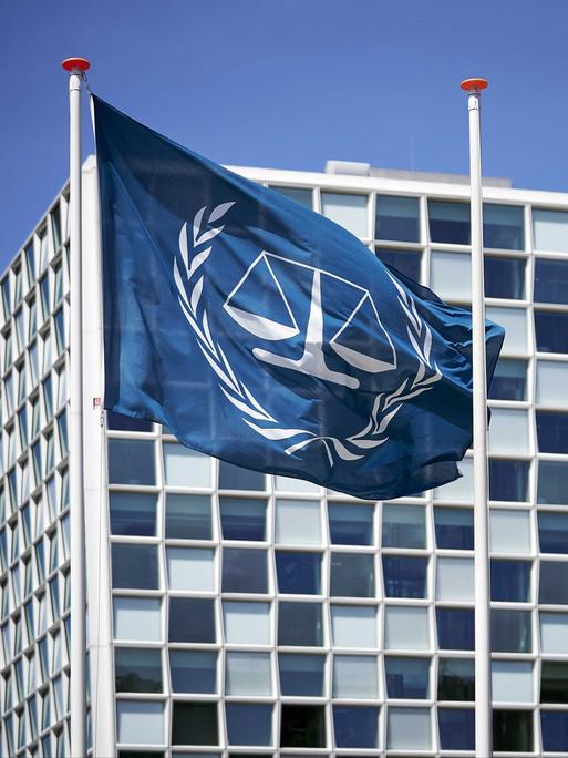 Außenansicht des Internationalen Strafgerichtshofs (IStGH). Davor weht die Flagge des IStGH, eine Waage mit zwei Waagschalen, die von zwei Olivenzweigen umschlossen wird.