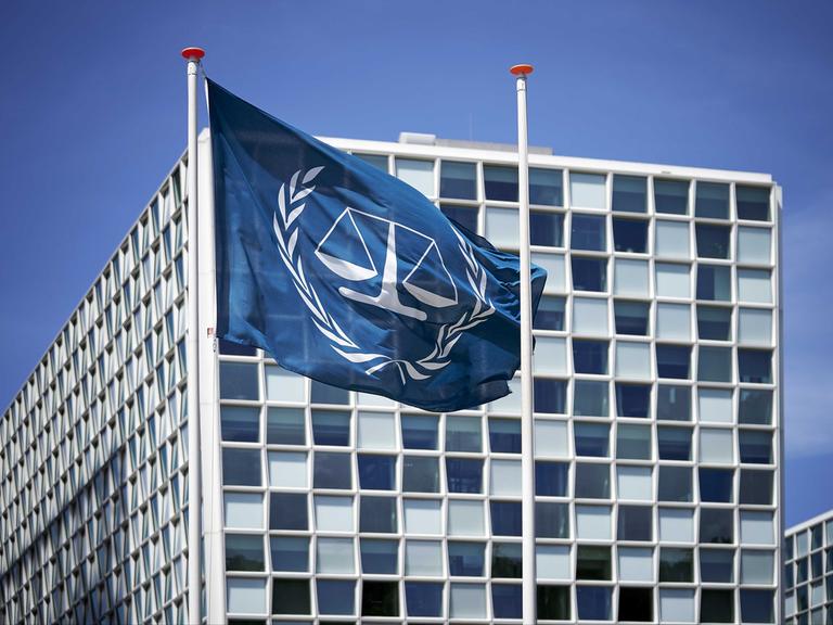 Außenansicht des Internationalen Strafgerichtshofs (IStGH). Davor weht die Flagge des IStGH, eine Waage mit zwei Waagschalen, die von zwei Olivenzweigen umschlossen wird.