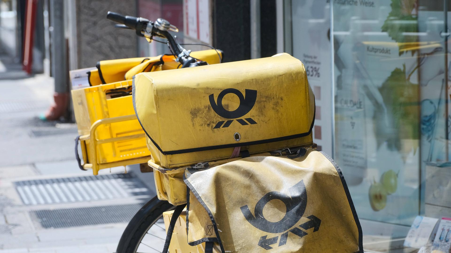 Zu sehen ist ein typisches, am Straßenrand abgestelltes gelbes Postfahrrad mit mehreren gelben Taschen und Boxen, in denen sich Zusendungen befinden.