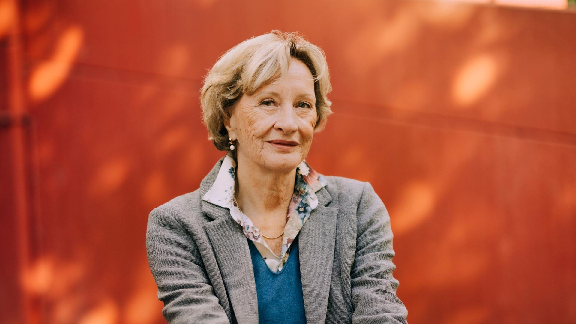 Helene Bracht sitzt mit übereinandergeschlagenen Beinen und schaut freundlich in die Kamera. Im Hintergrund ist ein Gebäude mit roter Fassade zu sehen. Helene Bracht sitzt mit übereinandergeschlagenen Beinen und schaut freundlich in die Kamera. Im Hintergrund ist ein Gebäude mit roter Fassade zu sehen.