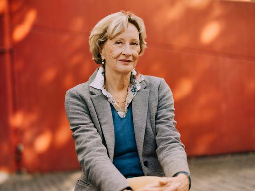 Helene Bracht sitzt mit übereinandergeschlagenen Beinen und schaut freundlich in die Kamera. Im Hintergrund ist ein Gebäude mit roter Fassade zu sehen. 