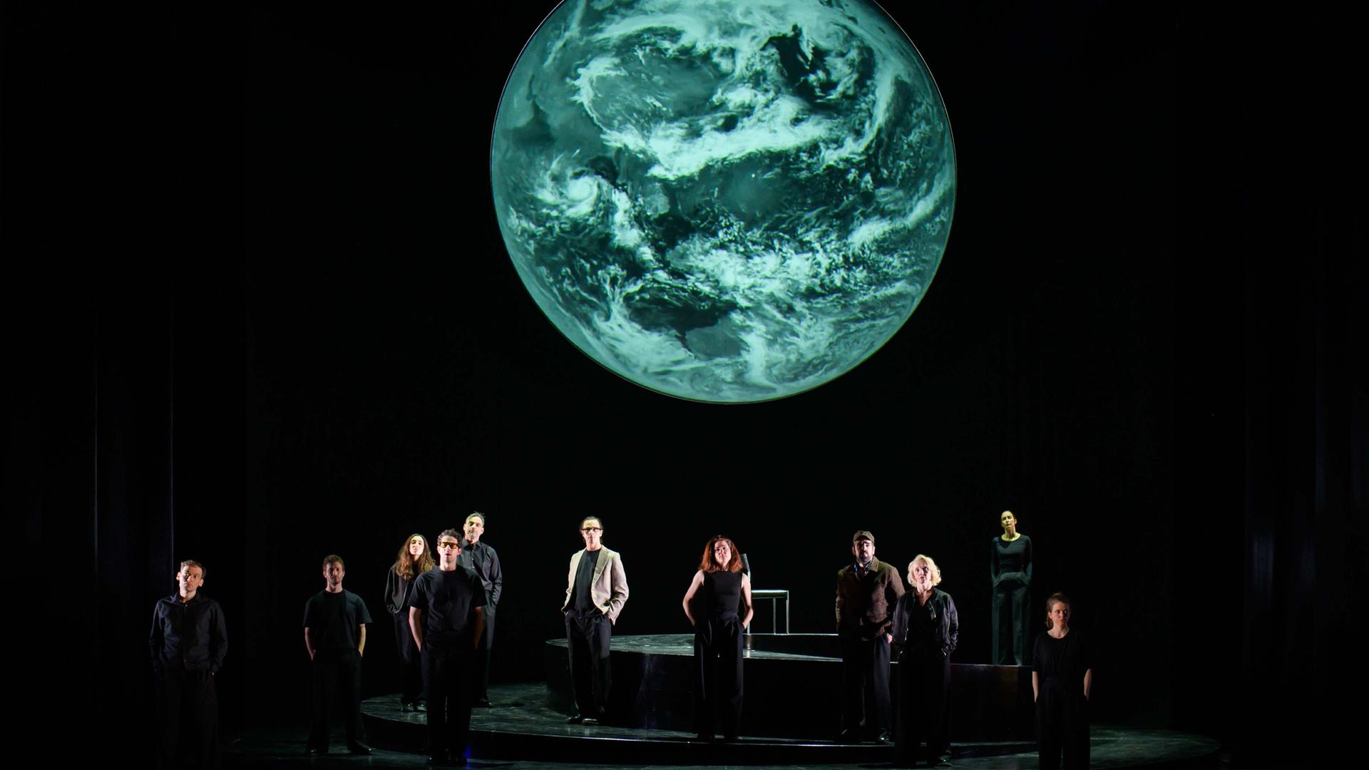 Szenenfoto aus der Produktion "Wir sind das Volk" am Deutschen Nationaltheater Weimar: Schauspielende auf einer dunklen Bühne blicken Richtung Publikum, hinter ihnen eine Projektion der Erdkugel. Szenenfoto aus der Produktion "Wir sind das Volk" am Deutschen Nationaltheater Weimar: Schauspielende auf einer dunklen Bühne blicken Richtung Publikum, hinter ihnen eine Projektion der Erdkugel.