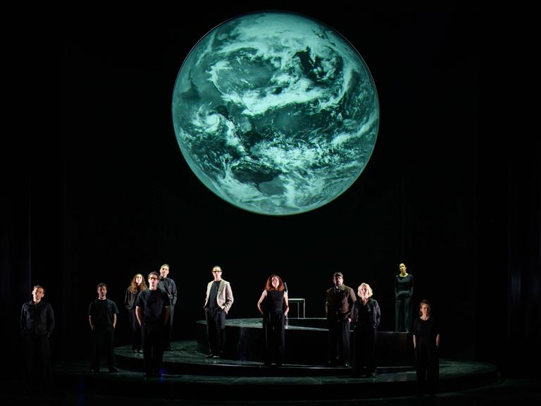 Szenenfoto aus der Produktion "Wir sind das Volk" am Deutschen Nationaltheater Weimar: Schauspielende auf einer dunklen Bühne blicken Richtung Publikum, hinter ihnen eine Projektion der Erdkugel. Szenenfoto aus der Produktion "Wir sind das Volk" am Deutschen Nationaltheater Weimar: Schauspielende auf einer dunklen Bühne blicken Richtung Publikum, hinter ihnen eine Projektion der Erdkugel.