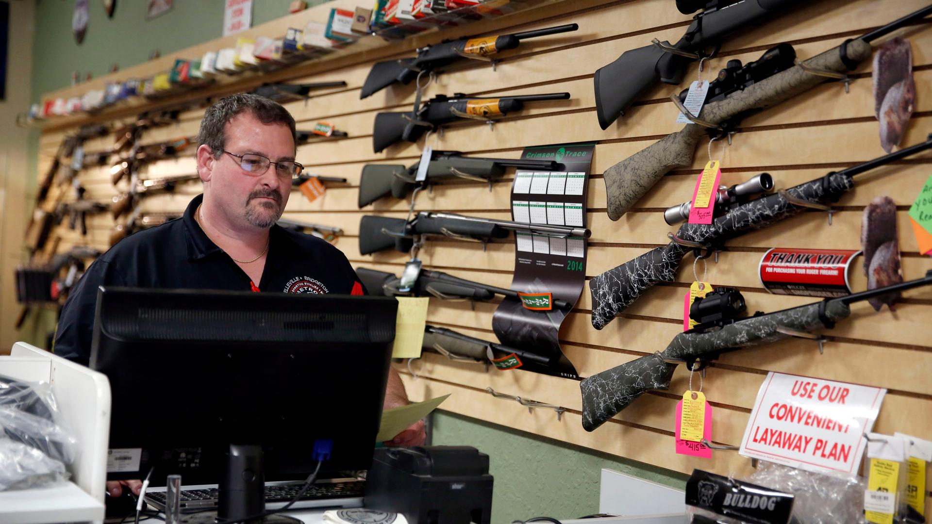 Eine Mann steht in einem US-Waffengeschäft hinter der Kasse. An der Wand hängen Dutzende Gewehre.