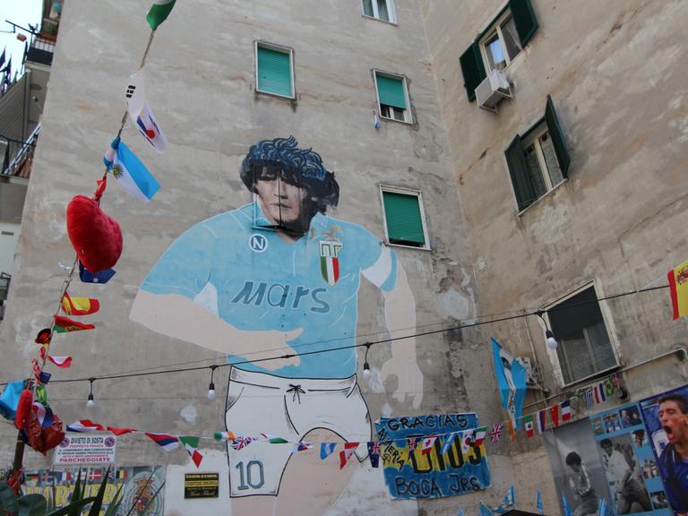 Auf einer Hauswand in Neapel prangt ein meterhohes Bild von Diego Maradona im Trikot des SSC Neapel. Der Fußballer hat schwarze, lockige Haare, das Trikot ist Blau und hat den Werbeschriftzug "Mars" auf der Brust.