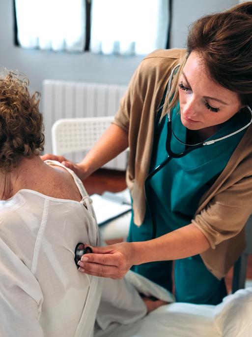 Eine Seniorin wird von einer jungen Pflegerin mit einem Stethoskop untersucht