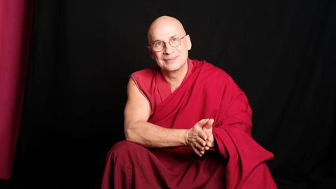 Tenzin Peljor sitzt in traditionell buddhistischem Gewand vor einem schwarzen Hintergrund und schaut freundlich in die Kamera.