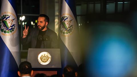 Nayib Bukele steht an einem Rednerpult und spricht zum Publikum. Links und rechts von ihm hängt die blau-weiße Flagge von El Salvador. Nayib Bukele steht an einem Rednerpult und spricht zum Publikum. Links und rechts von ihm hängt die blau-weiße Flagge von El Salvador.